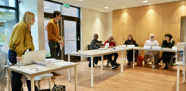 workshopleider Carin geeft workshop en staat voor een aantal bezoekers die aan tafel zitten