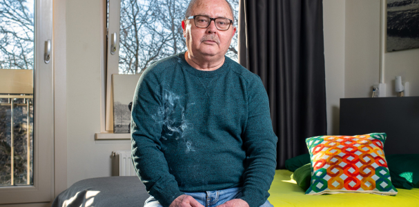 Meneer zit in een kamer op een kruk en kijkt in de camera.