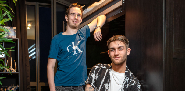 Twee jongens, een zit op een kruk en de ander staat met zijn arm tegen de muur aan 