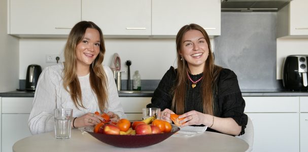 Twee dames lachen in de camera en zitten aan tafel met een schaal fruit