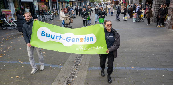 Een man en een vrouw lopen met een vlag waarop Buurt-Genoten staat.