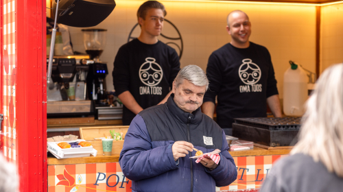 Twee mannen in een poffertjeskraam met een man die poffertjes eet daarvoor
