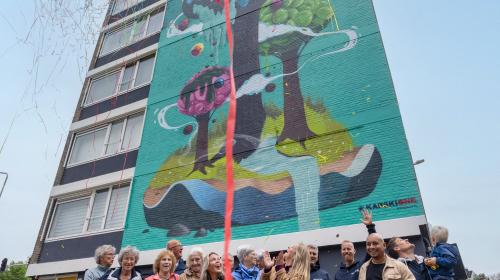 Feestelijke onthulling van de mural. Groepsfoto met lang rood lint van boven naar beneden. 