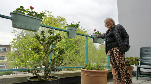 Mevrouw staat bij de rand van het balkon en laat haar bloemen zien, die hangen aan het hek.