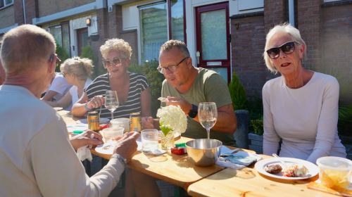 Bewoners zitten aan lange tafel in het zonnetje te genieten van het buurtfeestje
