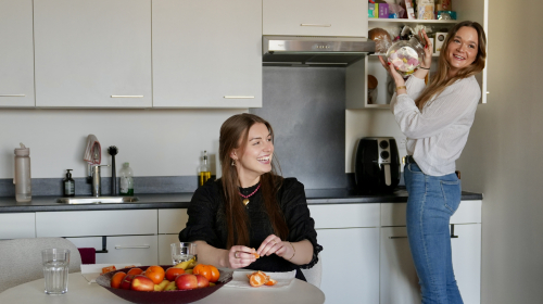 Twee dames in de keuken. Een aan tafel pelt een mandarijn en de andere pakt een pot uit de kast.