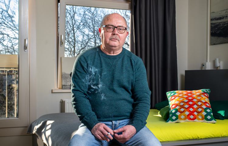 Meneer zit in een kamer op een kruk en kijkt in de camera.
