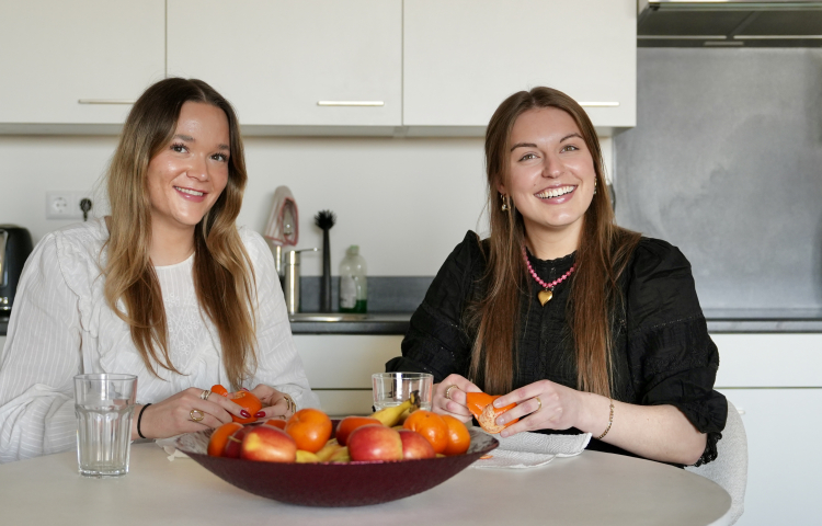 Twee dames lachen in de camera en zitten aan tafel met een schaal fruit