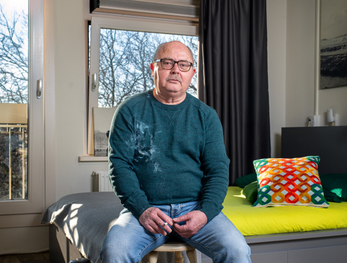 Meneer zit in een kamer op een kruk en kijkt in de camera.