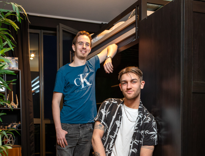 Twee jongens, een zit op een kruk en de ander staat met zijn arm tegen de muur aan 