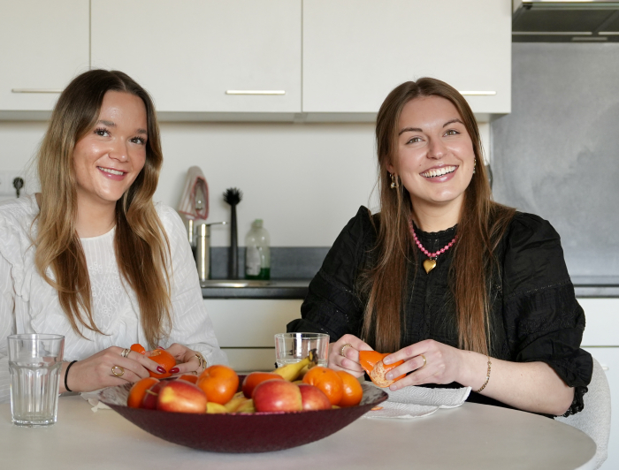 Twee dames lachen in de camera en zitten aan tafel met een schaal fruit